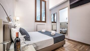 Una habitación de hotel moderna con una cama, una mesita de noche con una botella y vasos, una ventana con persianas, un espejo y un baño visible a través de una puerta abierta.