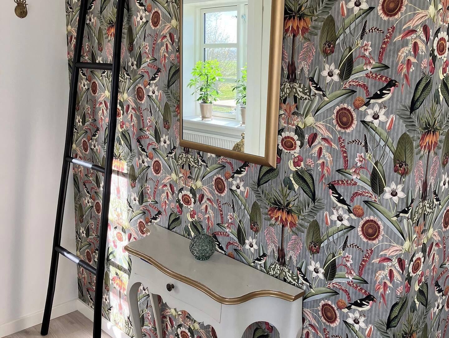 Una habitación con papel tapiz floral, un aparador, un espejo y una ventana con una planta.