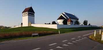 Ein Gebäude mit einem markanten Turm und ein großes Haus mit einem steilen Dach, auf einem grasbewachsenen Hügel neben einer Straße gelegen.