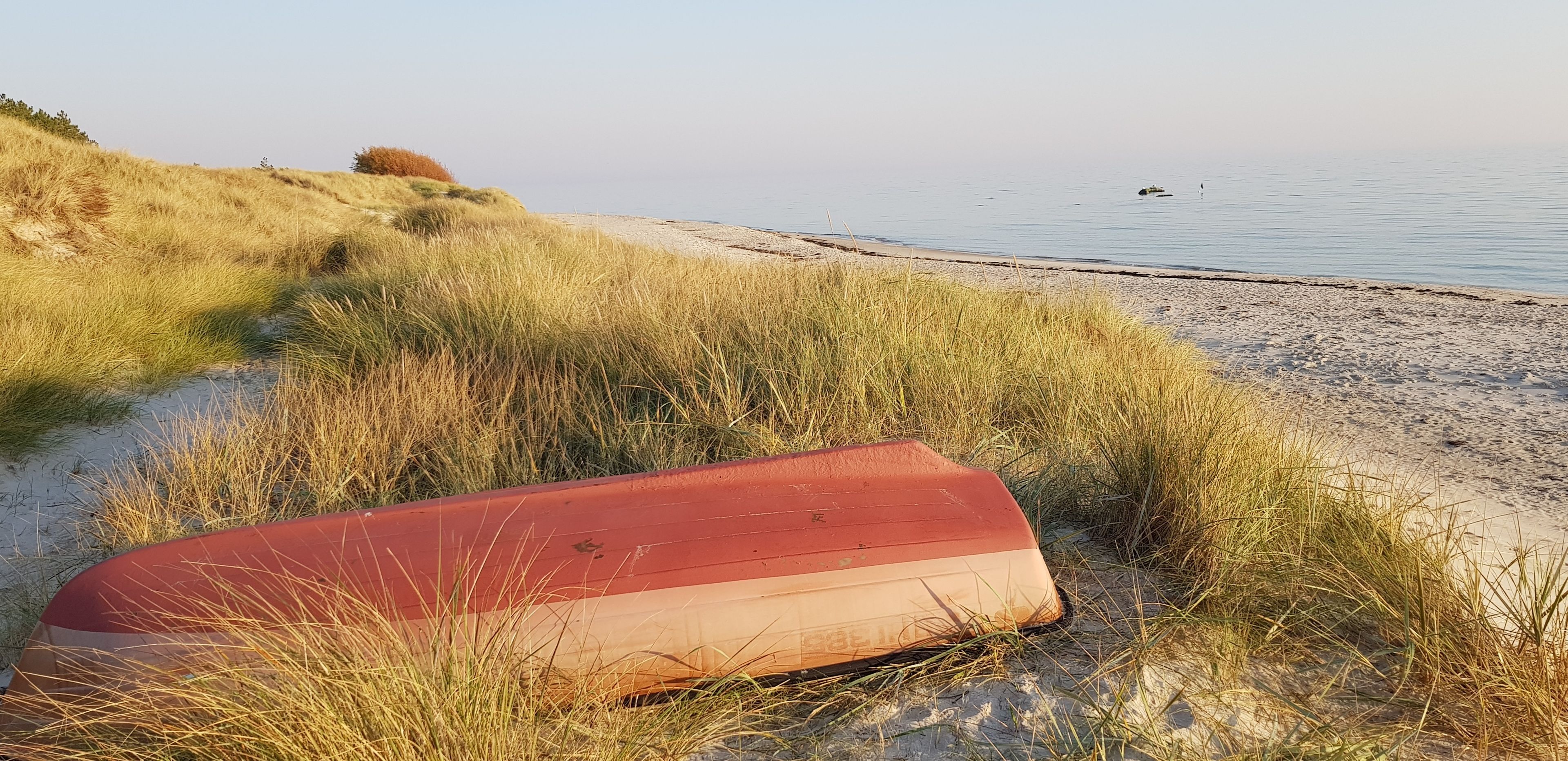 A beach with a red and beige boat lying on the sand, surrounded by grass.