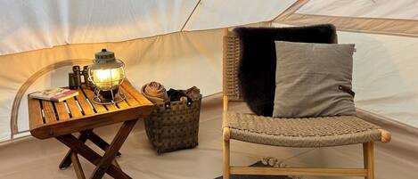 A tent interior with a wooden table, a lantern, a woven chair, and a grey cushion.