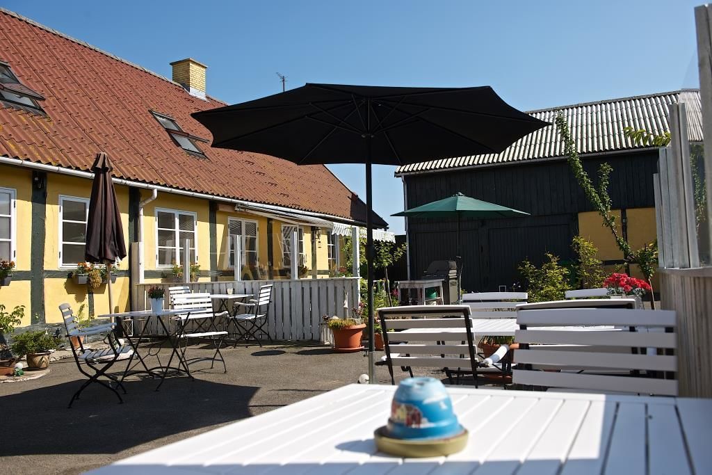 Un espace extérieur aménagé avec des tables et des chaises, un grand parasol, et un bâtiment doté d’un toit rouge et de murs jaunes.
