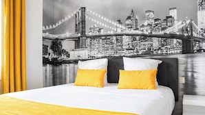Una cama con ropa blanca y dos almohadas amarillas, un mural con vista a la ciudad en la pared, y una cortina amarilla.