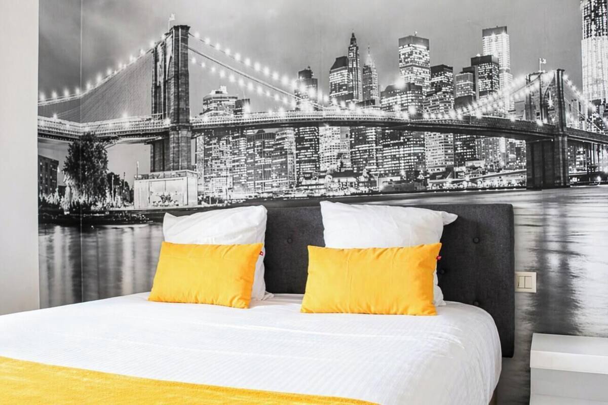 A bed with white bedding and two yellow pillows, a cityscape mural on the wall, and a yellow curtain.