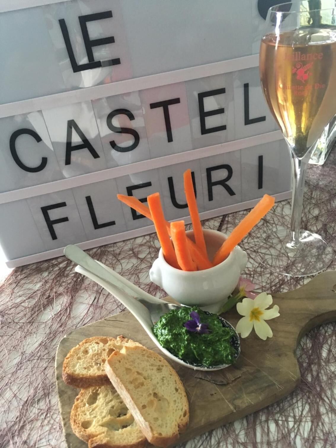 En dukat bord med en skylt som lyder ”LE CASTEL FLEURI”, ett glas vin, en skål med morotsstavar, en sked med grön dipp och brödskivor.