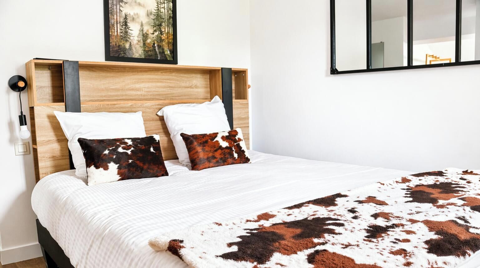 Un lit recouvert d’une couverture en peau de vache, deux oreillers à motifs de peau de vache, une tête de lit en bois et un tableau encadré au-dessus du lit.