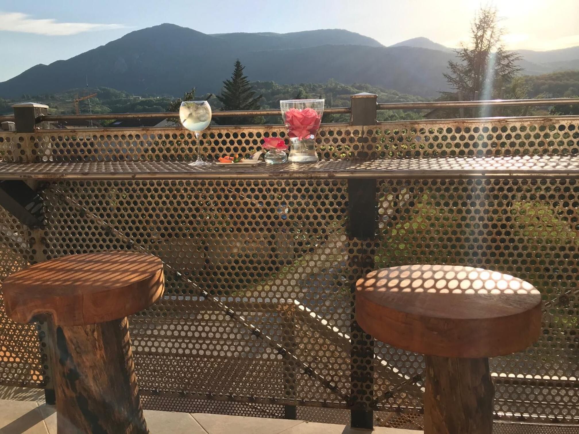 A table with a view of mountains, two stools, and a glass with a flower.