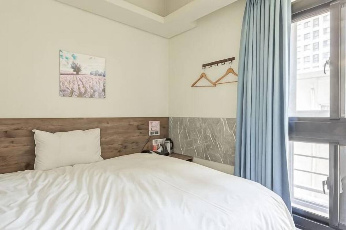 A hotel room with a bed, a nightstand, a window with curtains, and a wall-mounted coat rack.