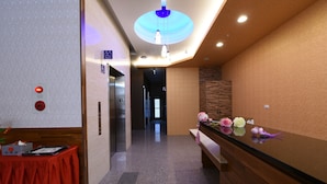 A hotel reception area with a wooden reception desk, a red table with a tissue box, and a bench. There is a hallway leading to an elevator and a restroom. - Hotel 99 (Taichung)