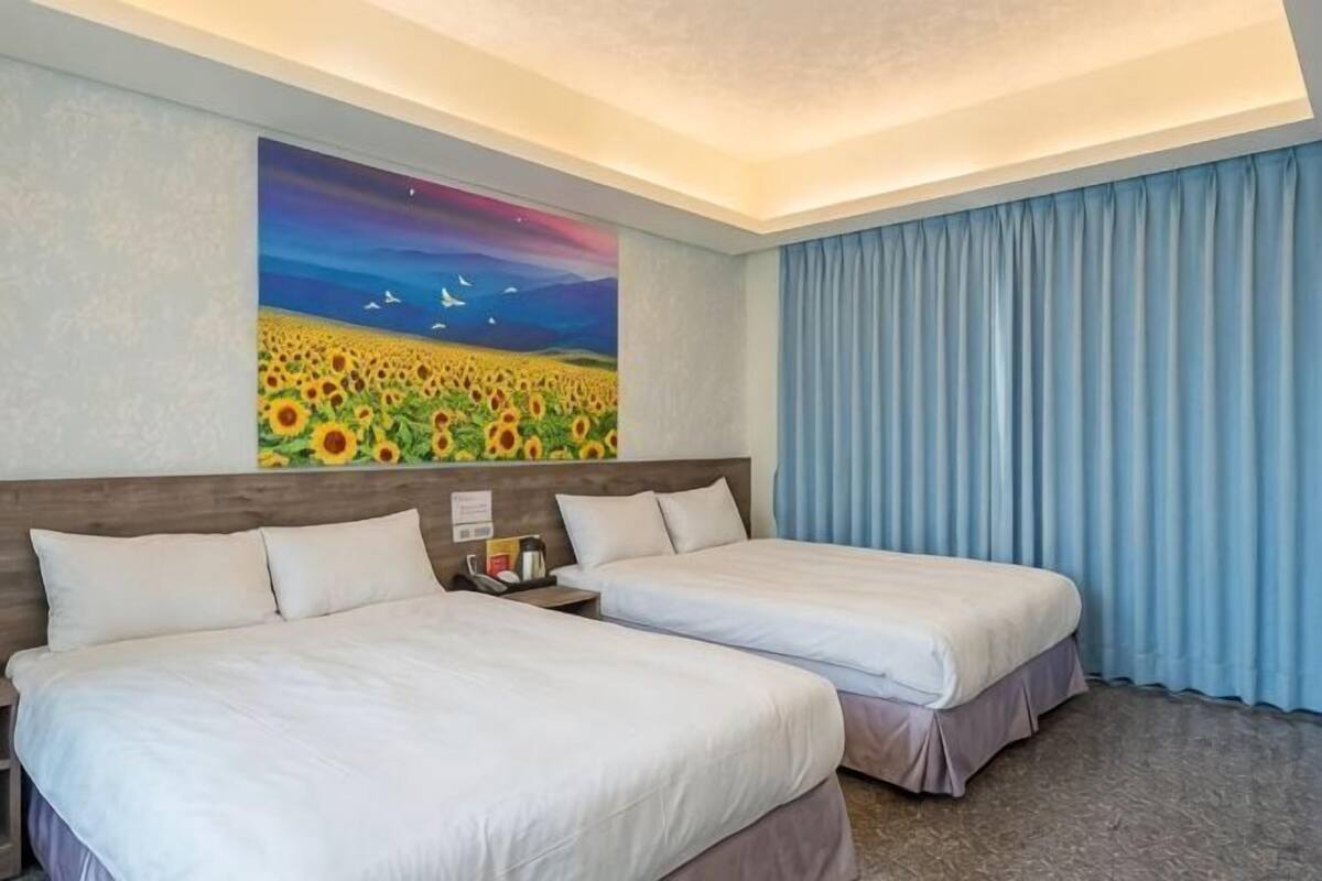 A hotel room with two beds, a painting of a sunflower field, and a blue curtain.