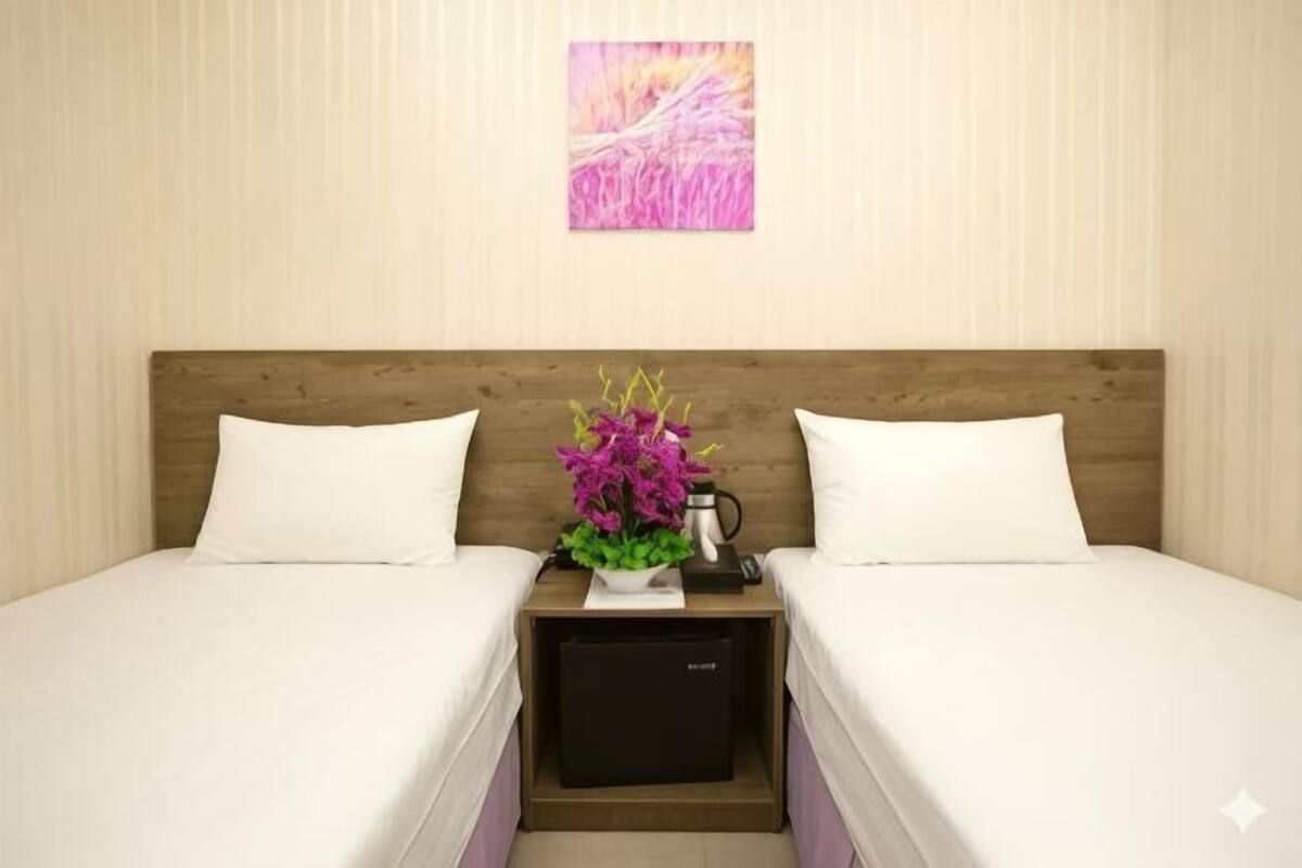 A hotel room with two beds, a wooden headboard, a nightstand with a vase of flowers, and a wall-mounted artwork.