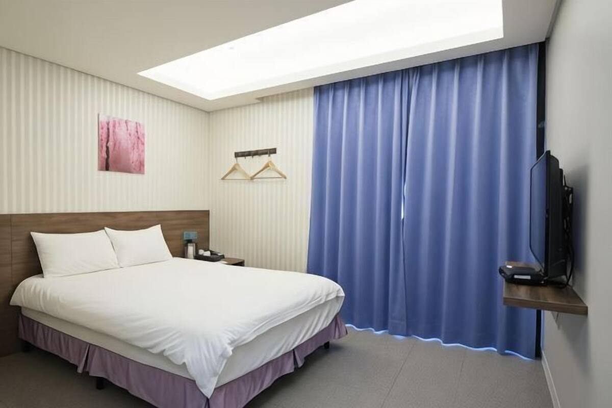 A hotel room with a bed, a TV, a nightstand, a window with curtains, and a wall-mounted light.