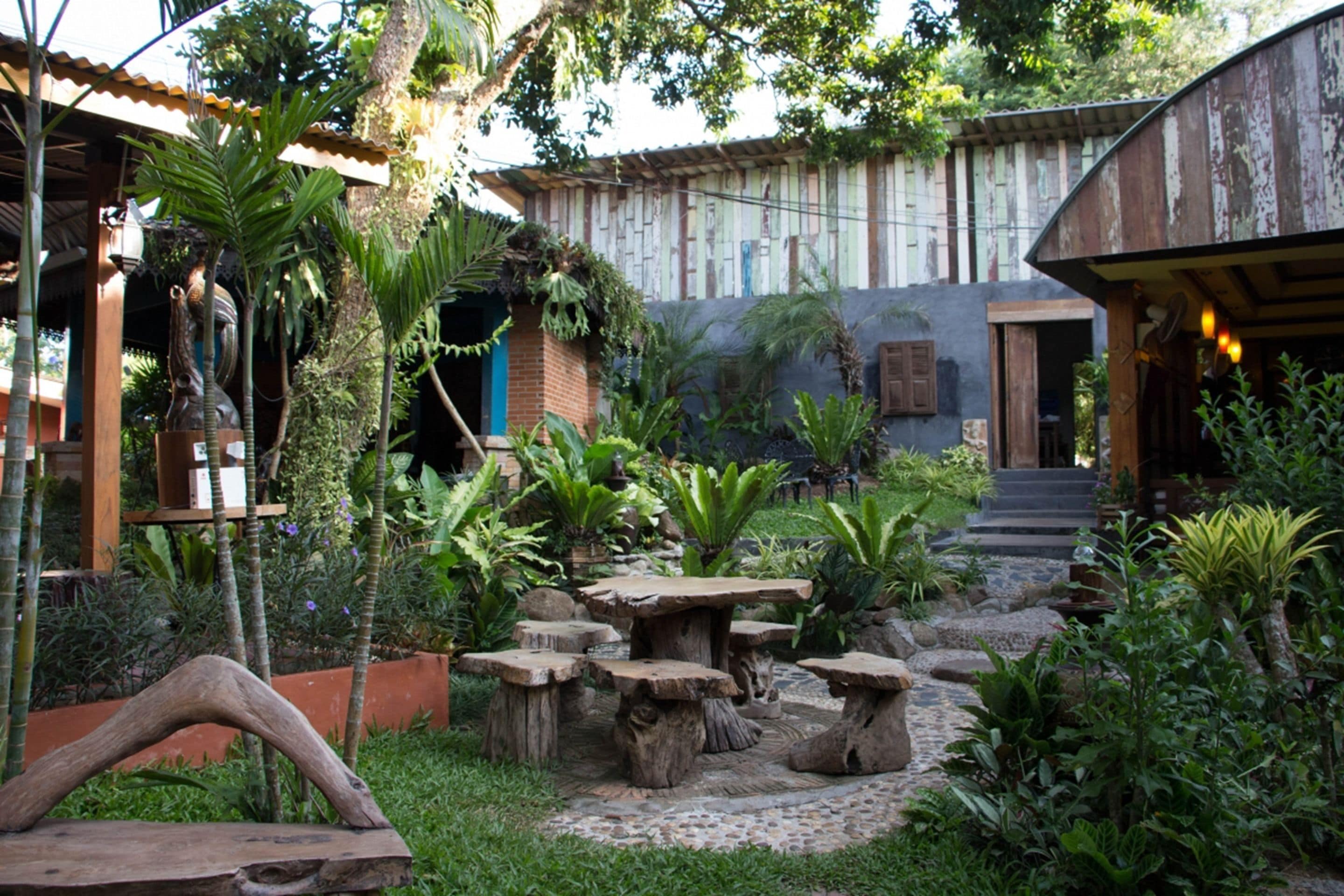 A rustic outdoor area with stone tables and benches, surrounded by lush greenery and a wooden structure.