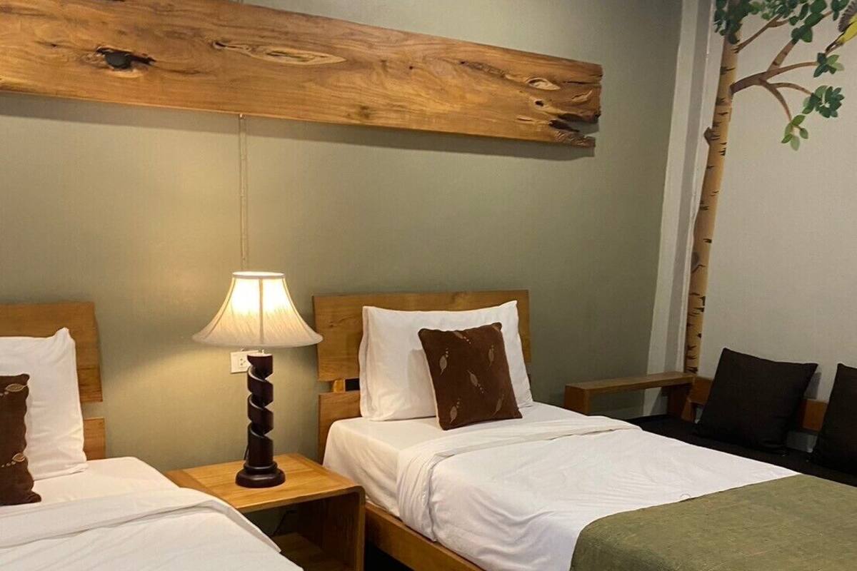 Habitación de hotel con dos camas, cabecera de madera, mesita de noche con lámpara y una manta verde sobre una de las camas.