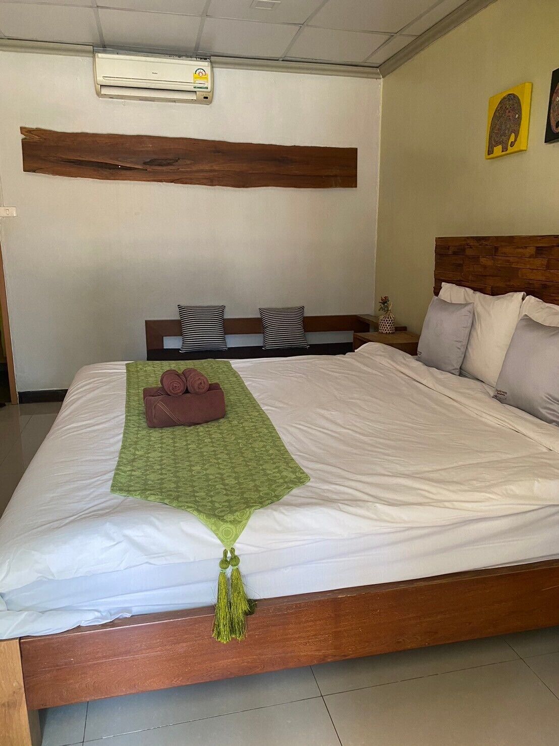 A neatly made bed with a green towel and a green tassel, wooden headboard, and a wall-mounted air conditioner.
