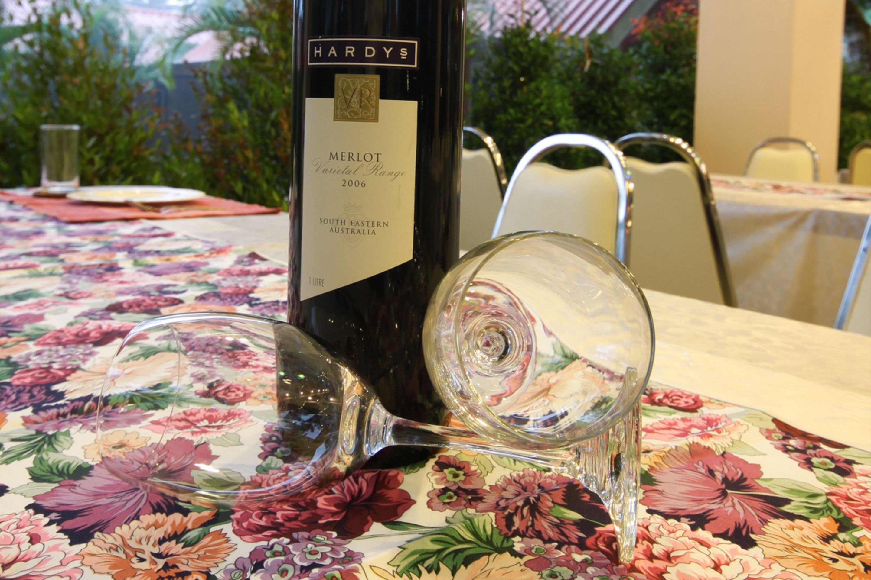 Una botella de vino Merlot y una copa de vino sobre una mesa con mantel floreado.