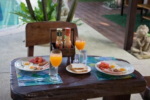 Une table en bois, dressée sur une terrasse avec vue sur une piscine et un jardin luxuriant, est garnie d’aliments pour le petit-déjeuner, notamment des œufs, des toasts et un verre de jus d’orange.