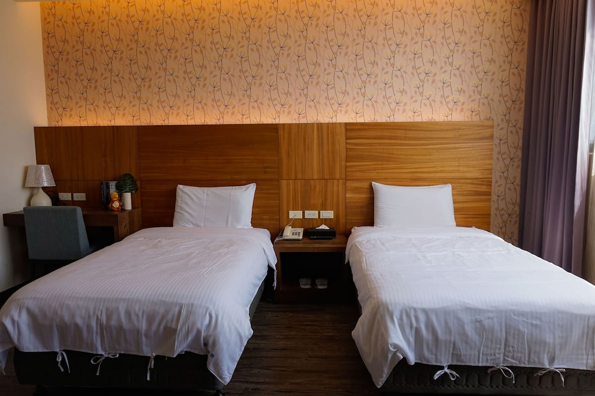 A hotel room with two single beds, wooden headboards, a nightstand with a lamp, a small table with a plant, and a patterned wallpaper.