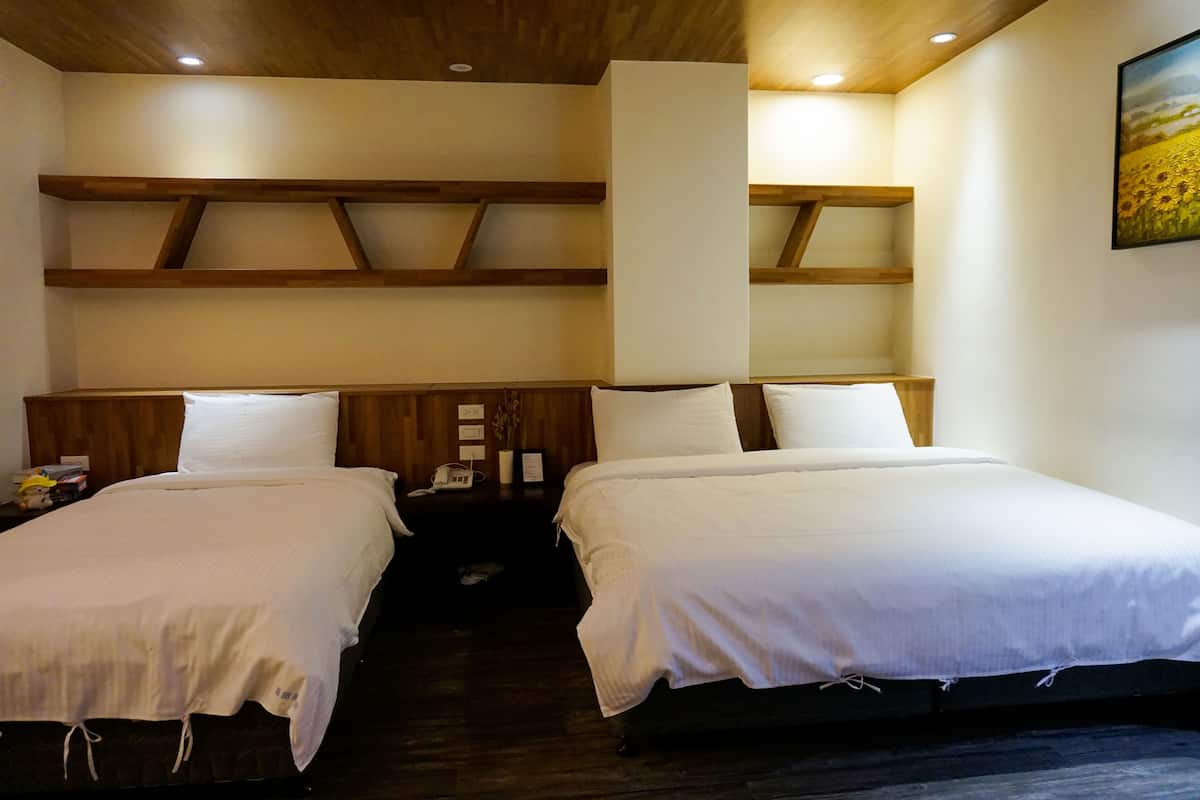 A hotel room with two beds, wooden headboards, a wall-mounted shelf, and a painting of a field.
