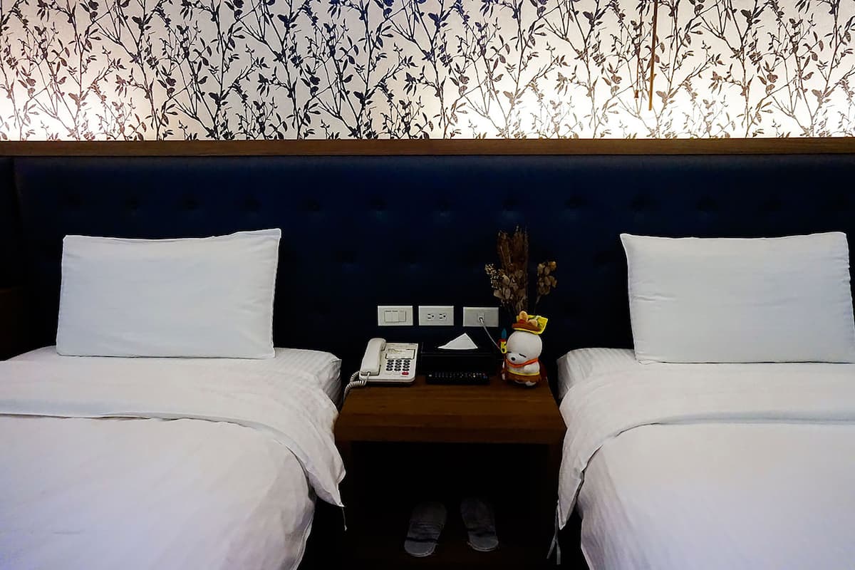 A hotel room with two beds, a nightstand with a phone, a vase with flowers, and a wall with a floral pattern.