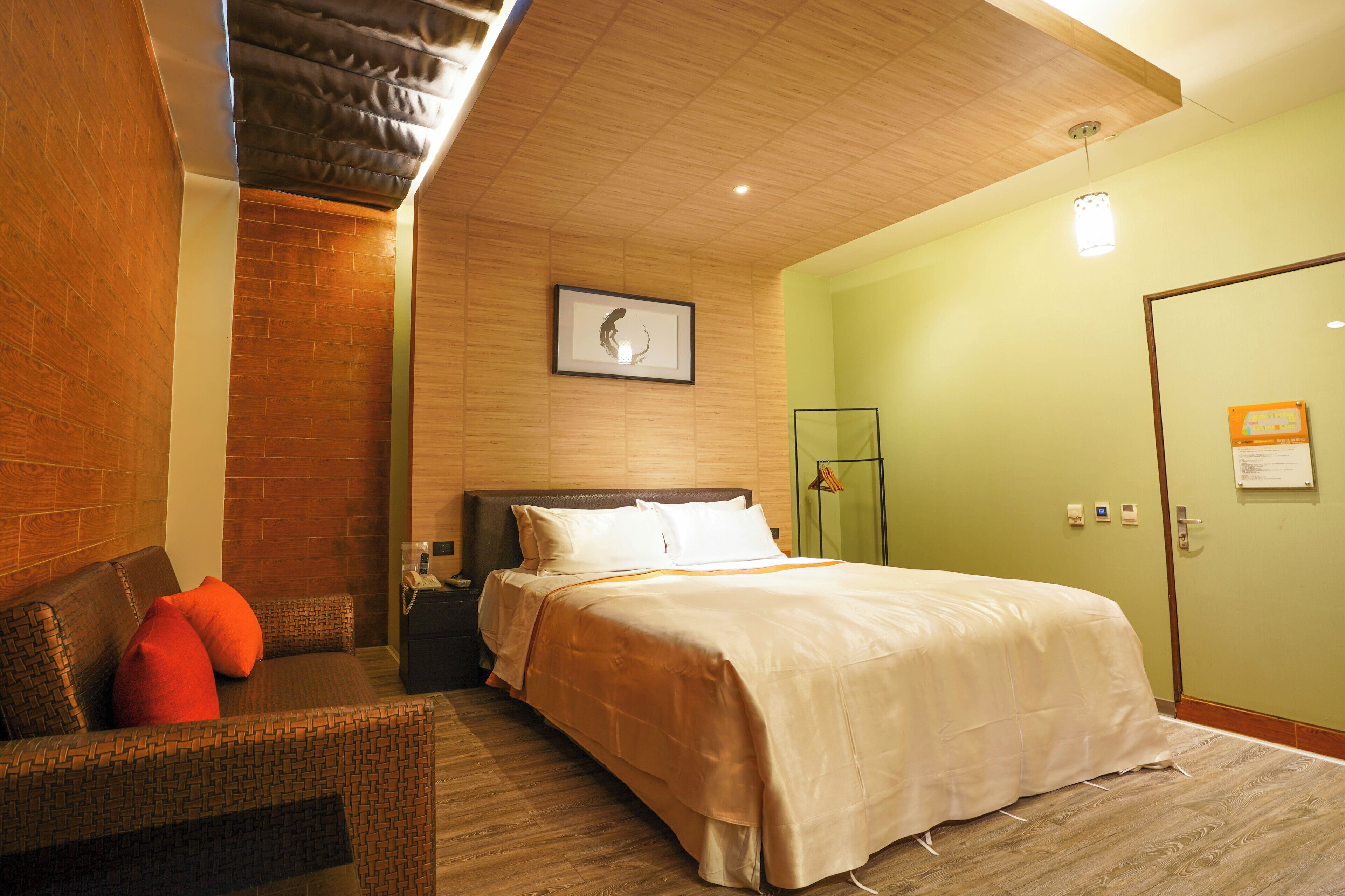 A hotel room with a bed, a nightstand, a seating area with a chair and cushions, a framed artwork on the wall, and a door with a sign.