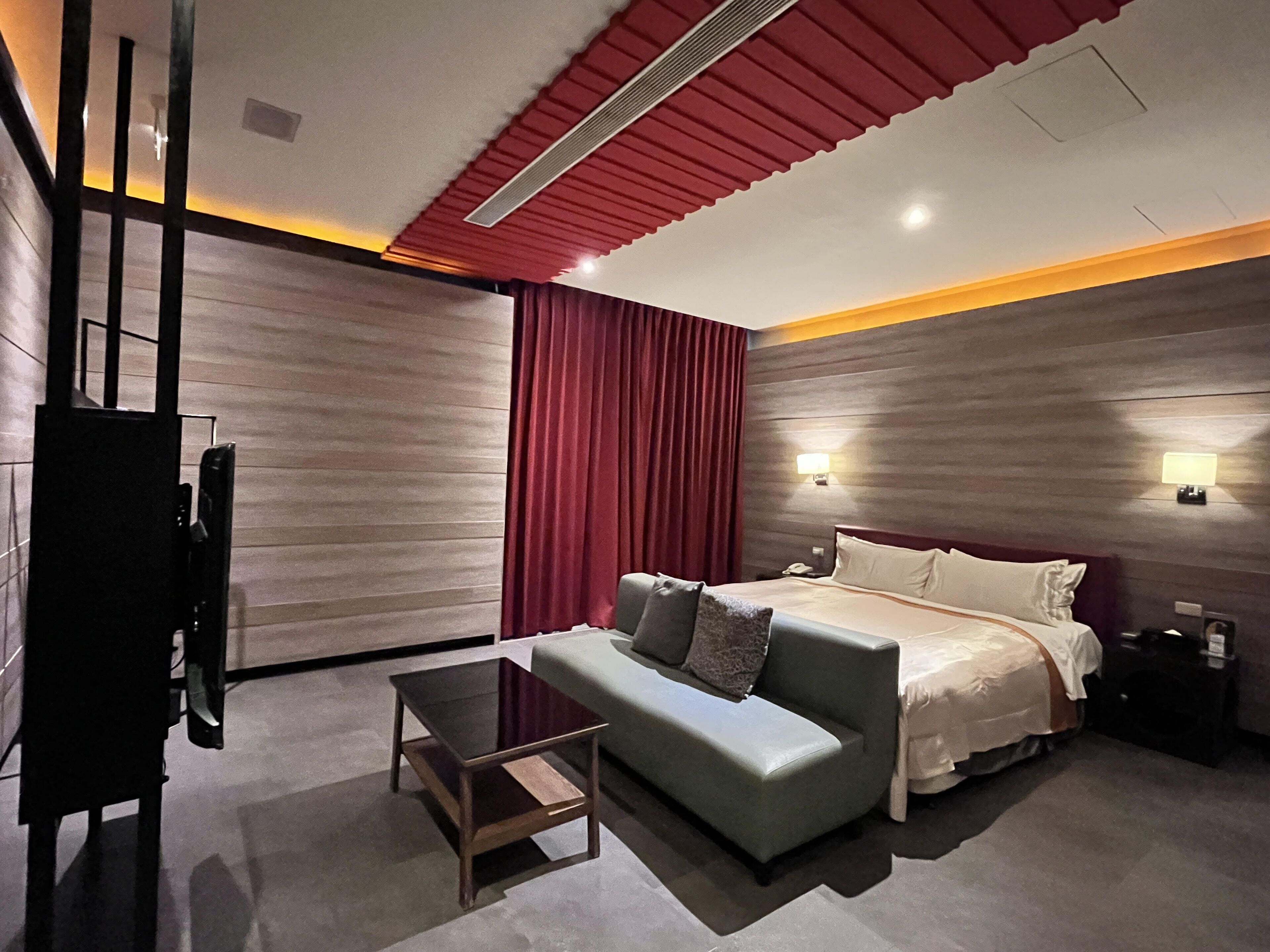 A modern hotel room with a bed, a sofa, a small table, a TV, and a red curtain.