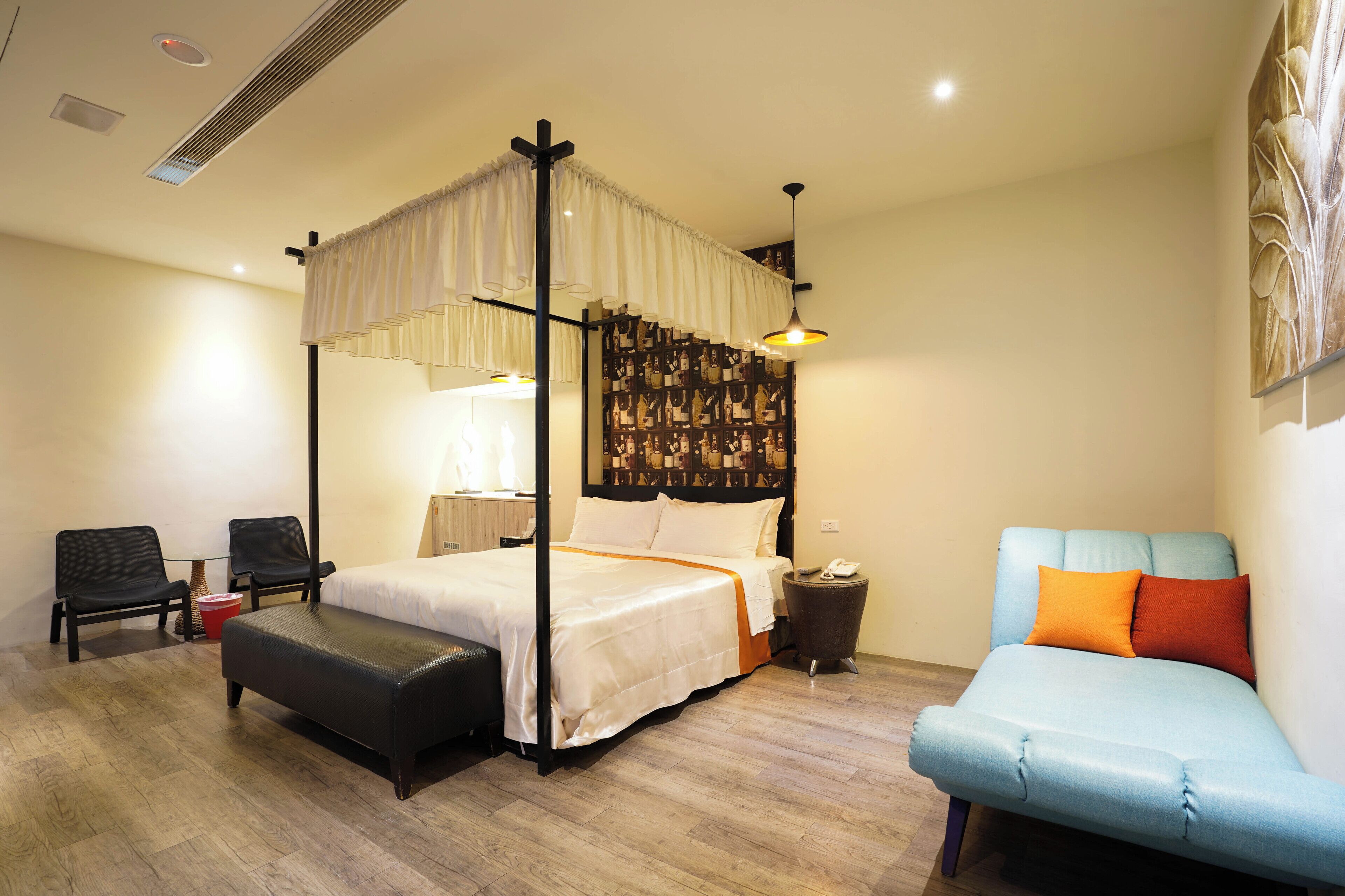 A four-poster bed with a canopy, a blue sofa with orange cushions, a small table with a lamp, and a wall with a patterned artwork.