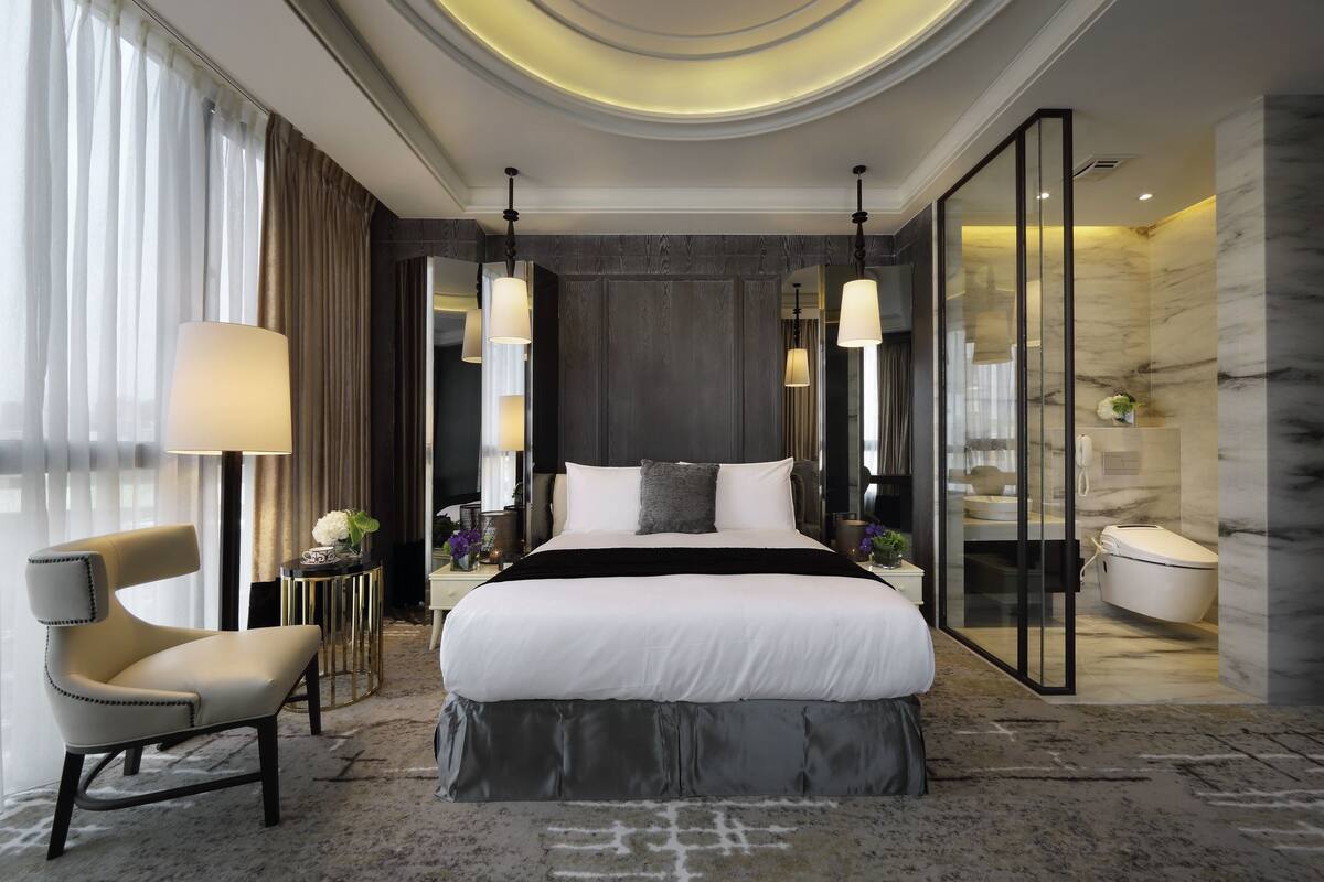 A modern hotel room with a large bed, a sitting area with a chair, a small table with a lamp, and a bathroom with a toilet and sink.