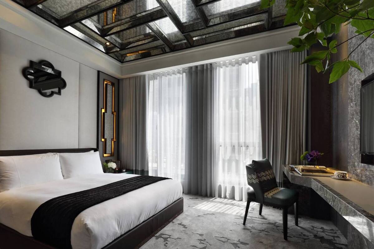 A modern hotel room with a large bed, a desk with a chair, and a large window with curtains.