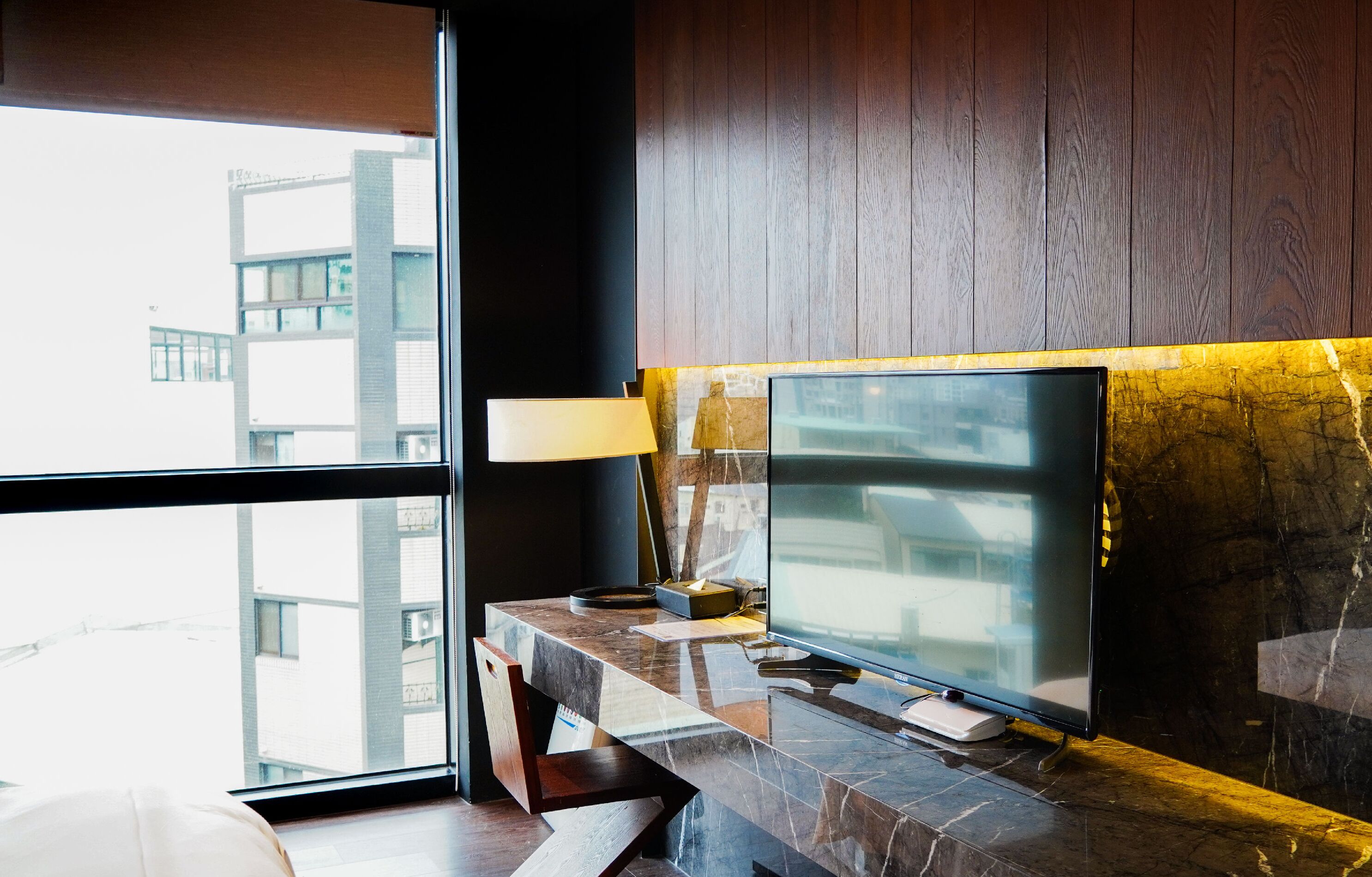 A modern hotel room with a large window, a flat-screen TV on a marble console, and a wooden wall.