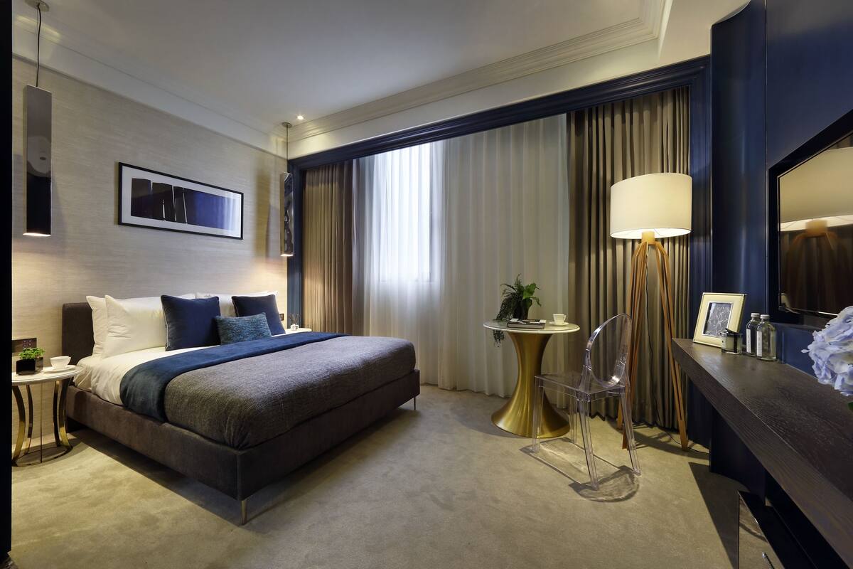 A modern hotel room with a large bed, bedside table, lamp, and a small round table with a chair.