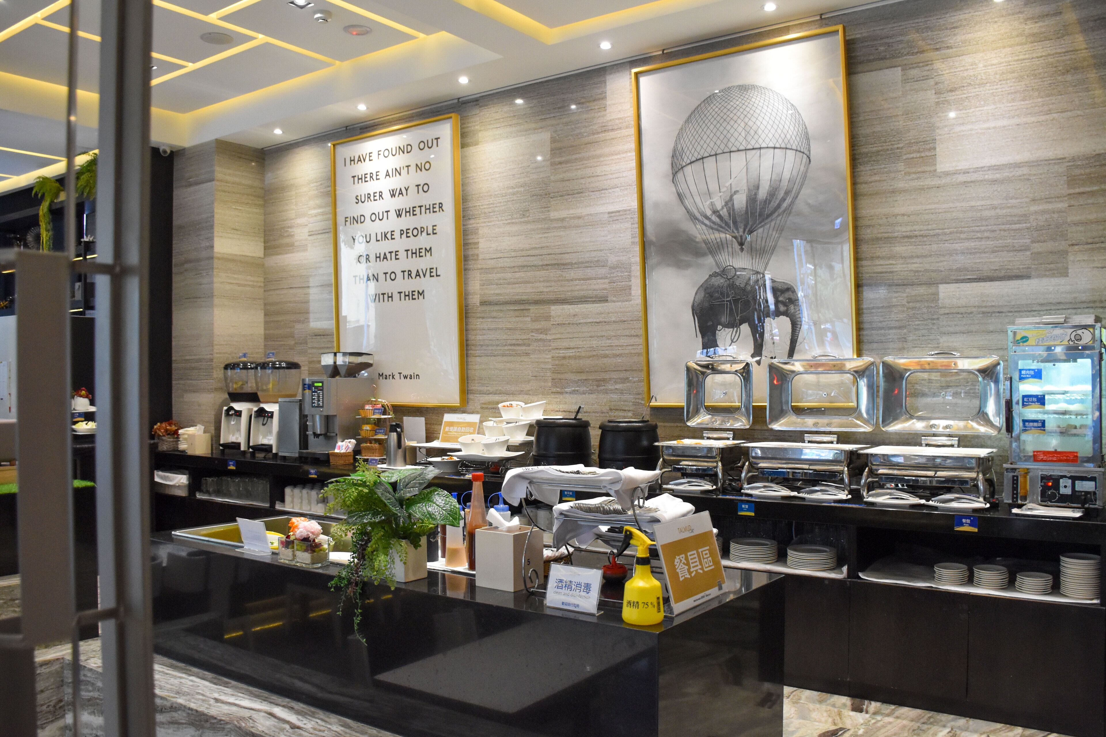 Zona de bufé con diferentes estaciones de comida, una máquina de café y una cita en un marco grande en la pared.
