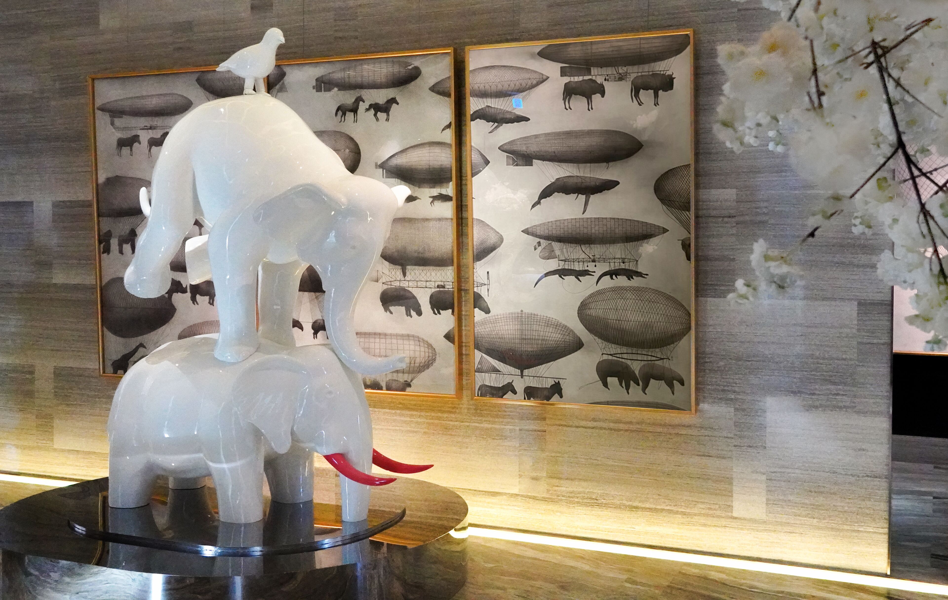 A sculpture of a white elephant with red tusks on a pedestal, set against a backdrop of framed artwork depicting various animals and objects.