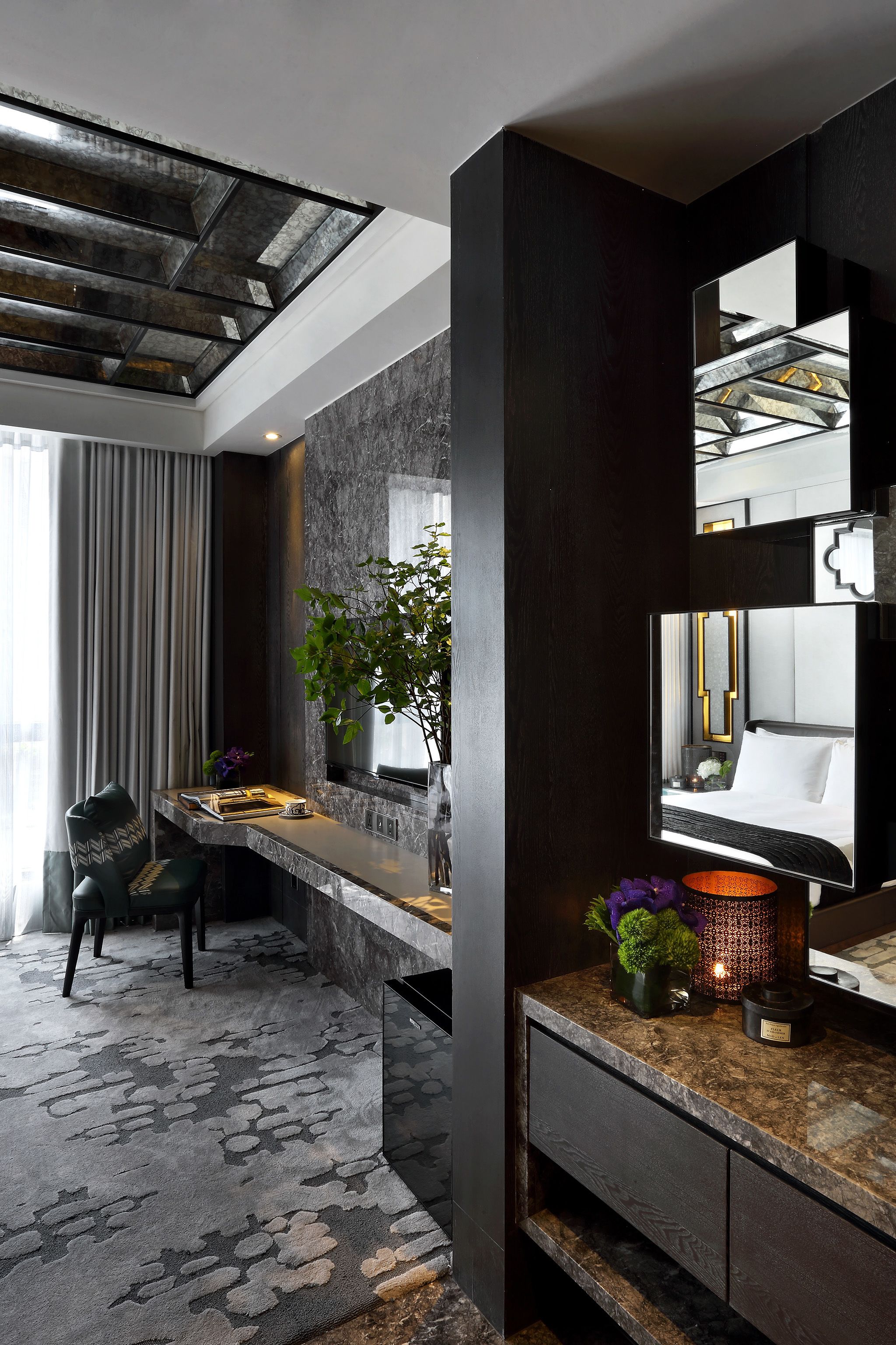 Une chambre d’hôtel moderne avec un décor en bois foncé, un grand miroir, un bureau agrémenté d’une plante et une vue sur la ville grâce à une verrière.