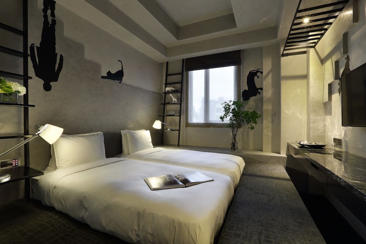 A modern hotel room with a large bed, bedside lamps, a nightstand, a window with a view, and a small plant.