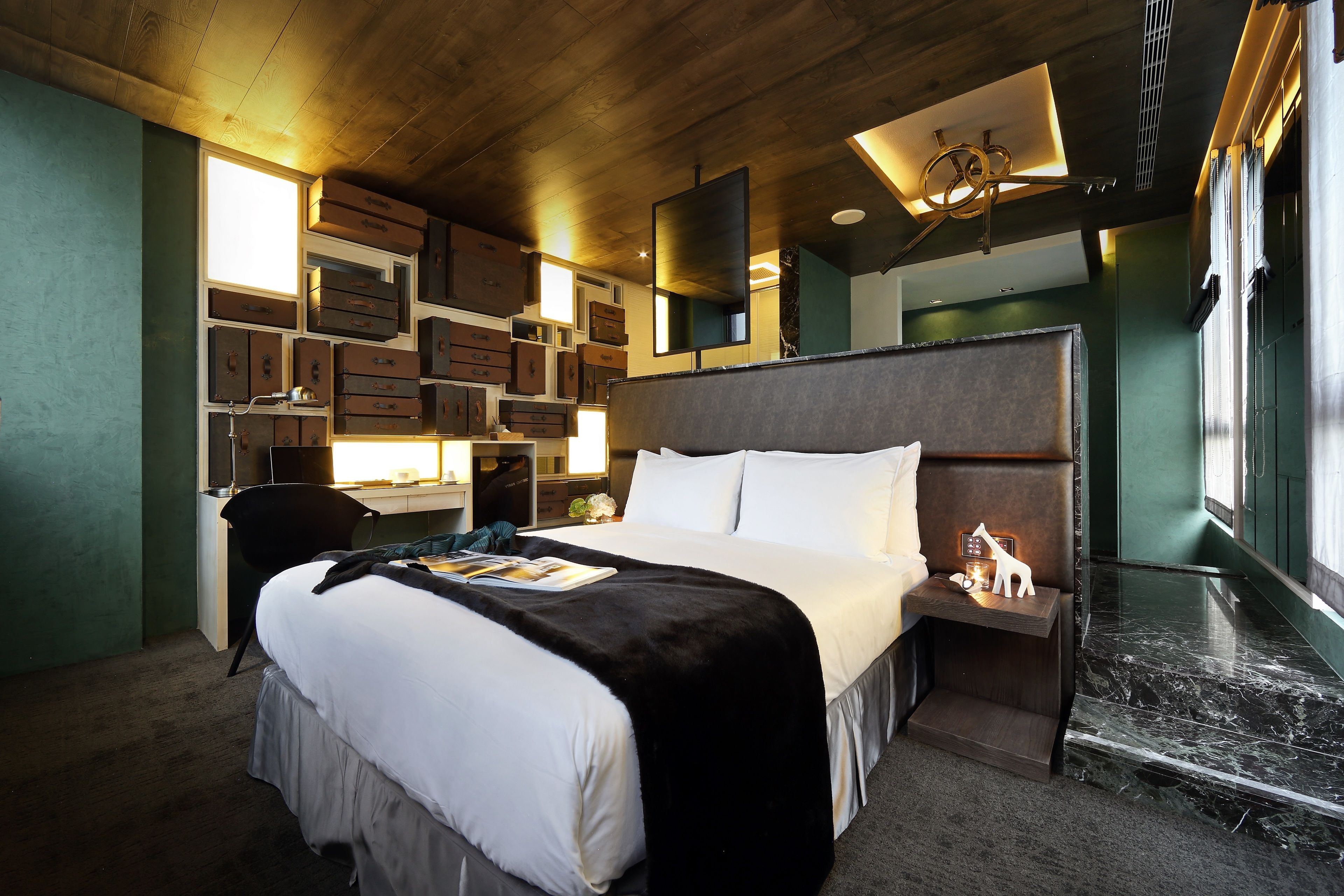 Habitación de hotel con una cama grande, un escritorio con cajones, una silla, una mesita de noche con lámpara y piso de mármol.