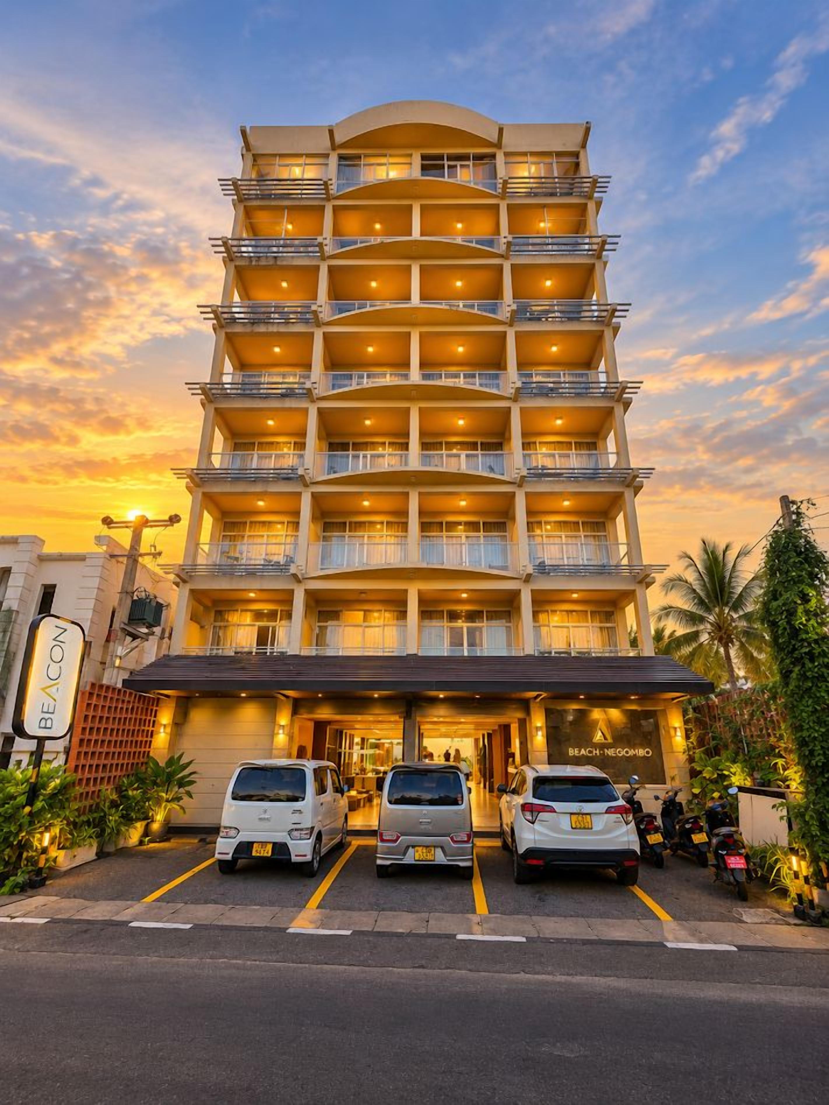 Un hôtel à plusieurs étages doté de balcons, une enseigne indiquant « BEACON » et une autre indiquant « BEACH NEIGOMBO ». Des voitures sont garées devant.