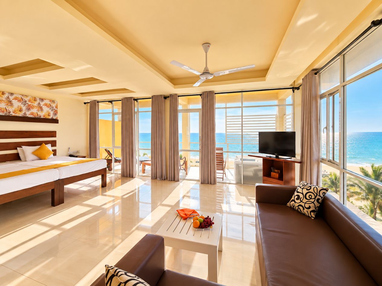 A spacious living room with a large window, a ceiling fan, a flat-screen TV, and a sofa with a view of the ocean.