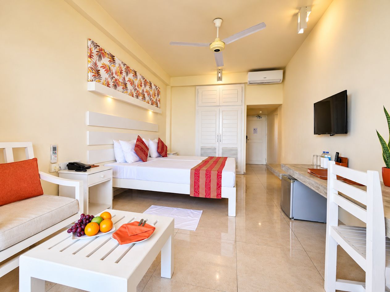 A hotel room with a bed, a white sofa, a dining table with fruit, a TV, and a ceiling fan.
