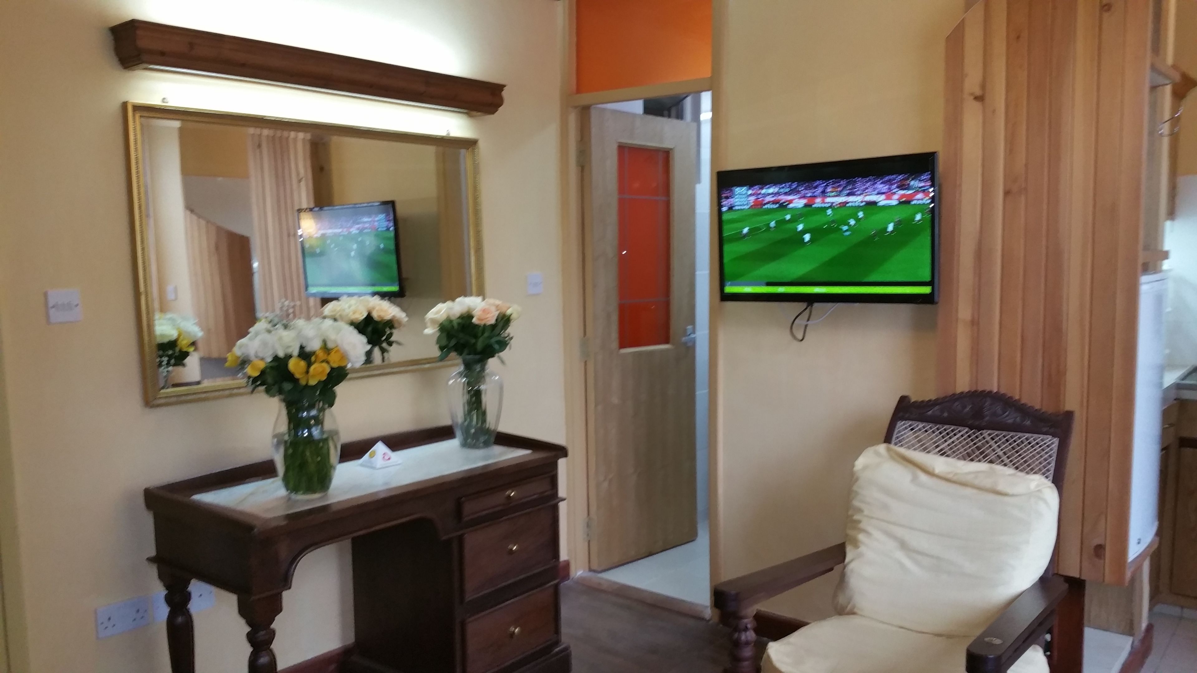 A room with a wooden desk, a chair, a large mirror, and a flat-screen TV mounted on the wall.