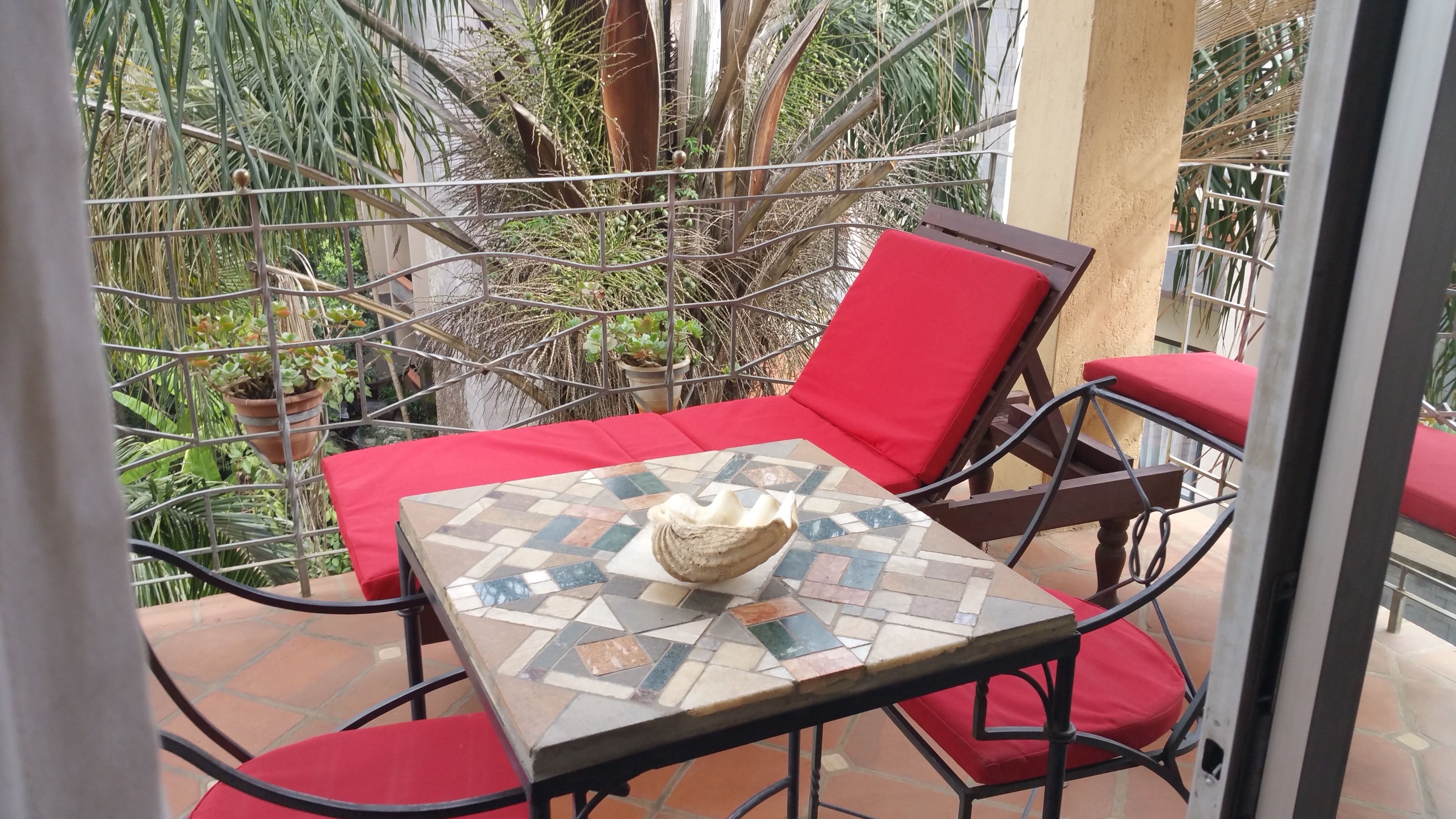 A small outdoor seating area with a tiled table and red cushioned chairs, surrounded by a metal fence and potted plants.