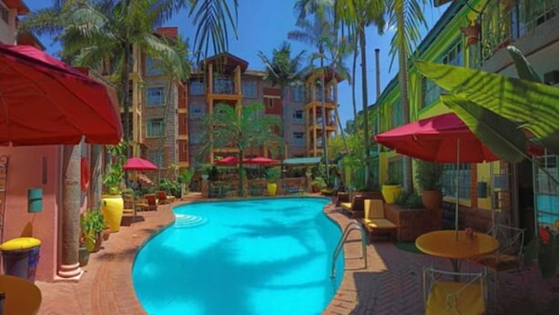 A swimming pool surrounded by palm trees and colorful umbrellas, with a multi-story building in the background. - Woodmere Serviced Apartments (Nairobi)