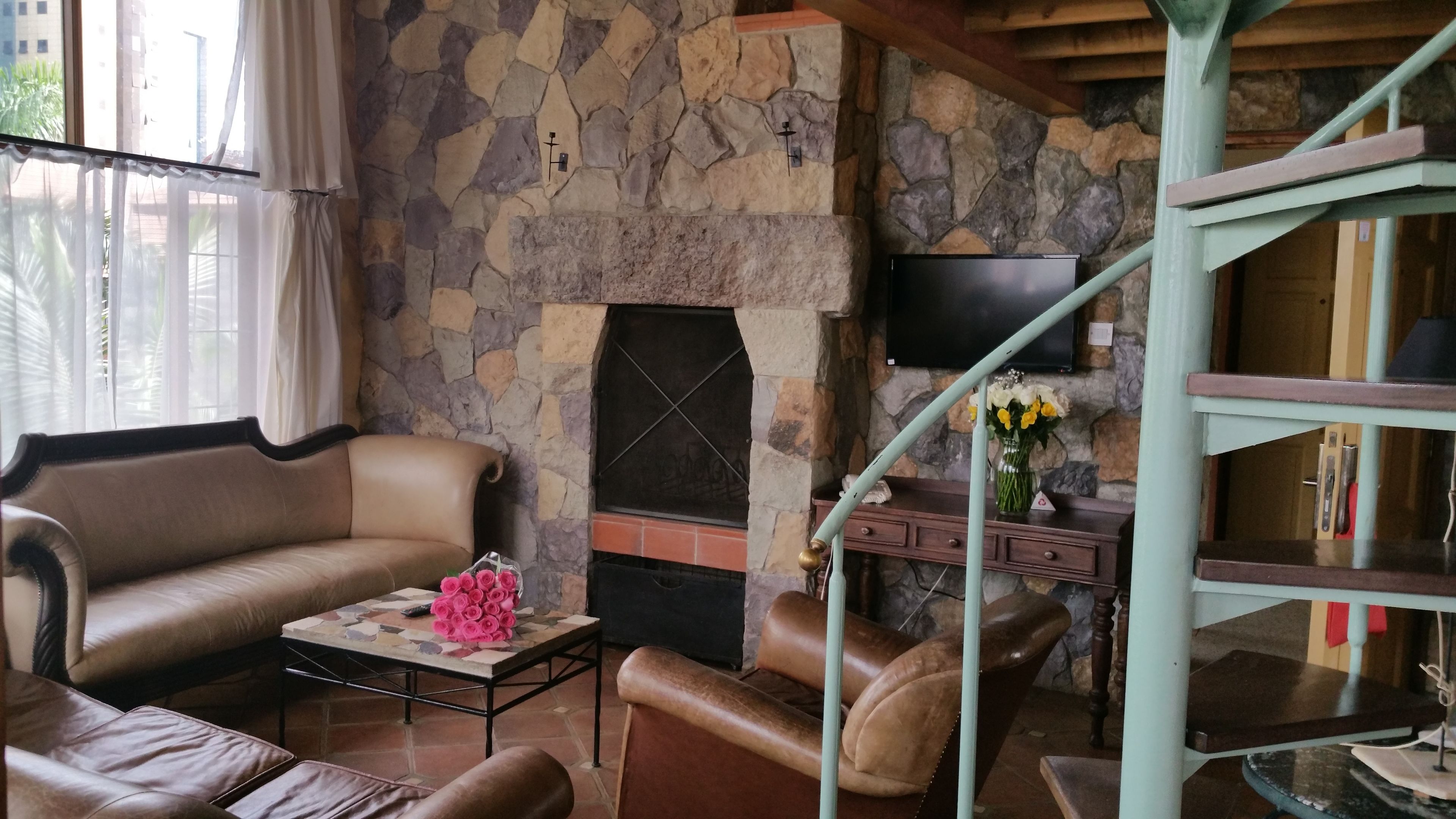 A cozy living room with a stone fireplace, leather sofas, a coffee table with a vase of flowers, and a TV mounted on the wall.