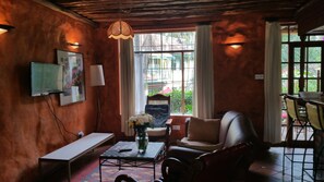 Une pièce chaleureuse avec un plafond en bois, une grande fenêtre donnant sur la verdure, un canapé, un fauteuil, une table et un téléviseur fixé au mur.
