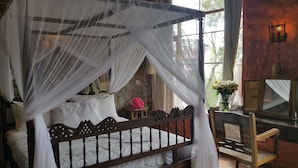 A four-poster bed with a stone wall backdrop, a small table with a pink object, and a chair with a woven seat. - Woodmere Serviced Apartments (Nairobi)