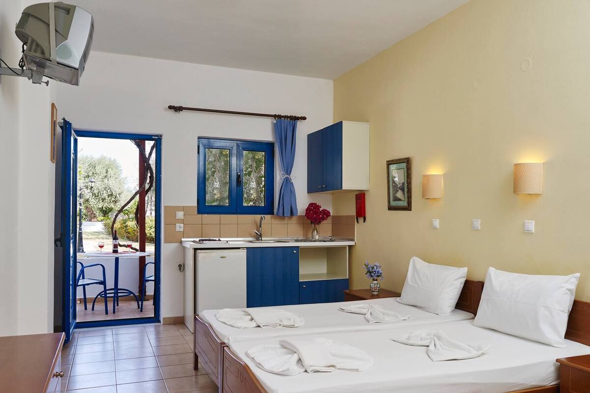 A compact hotel room with a bed, a small kitchenette, a dining area, and a view of the outdoors.