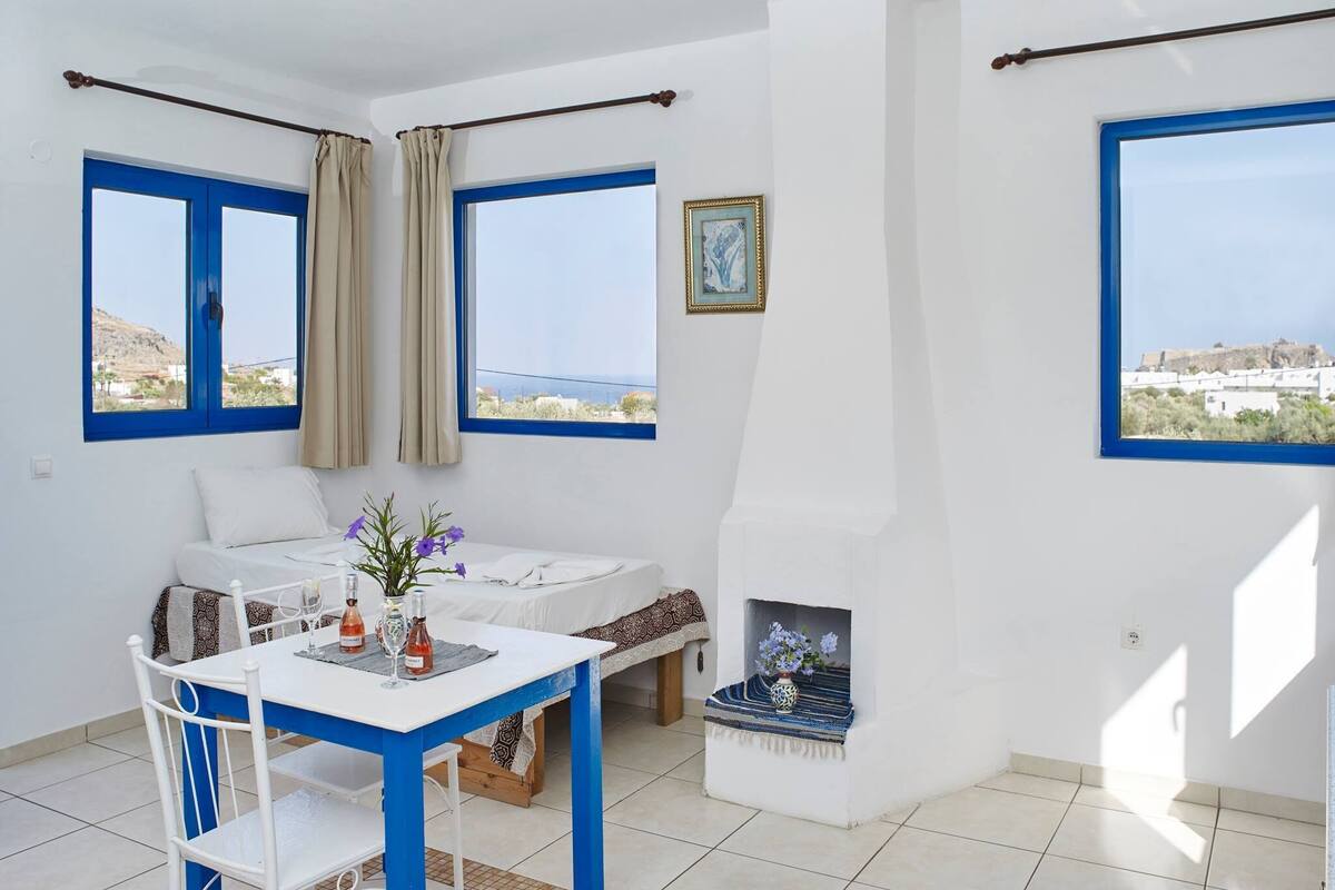 A room with a bed, a table with chairs, a fireplace, and large windows with blue frames.