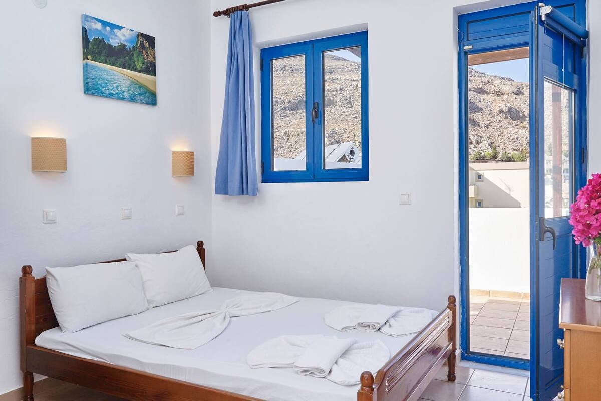 A bedroom with a wooden bed, a window with blue curtains, a wall-mounted lamp, and a painting of a beach scene.