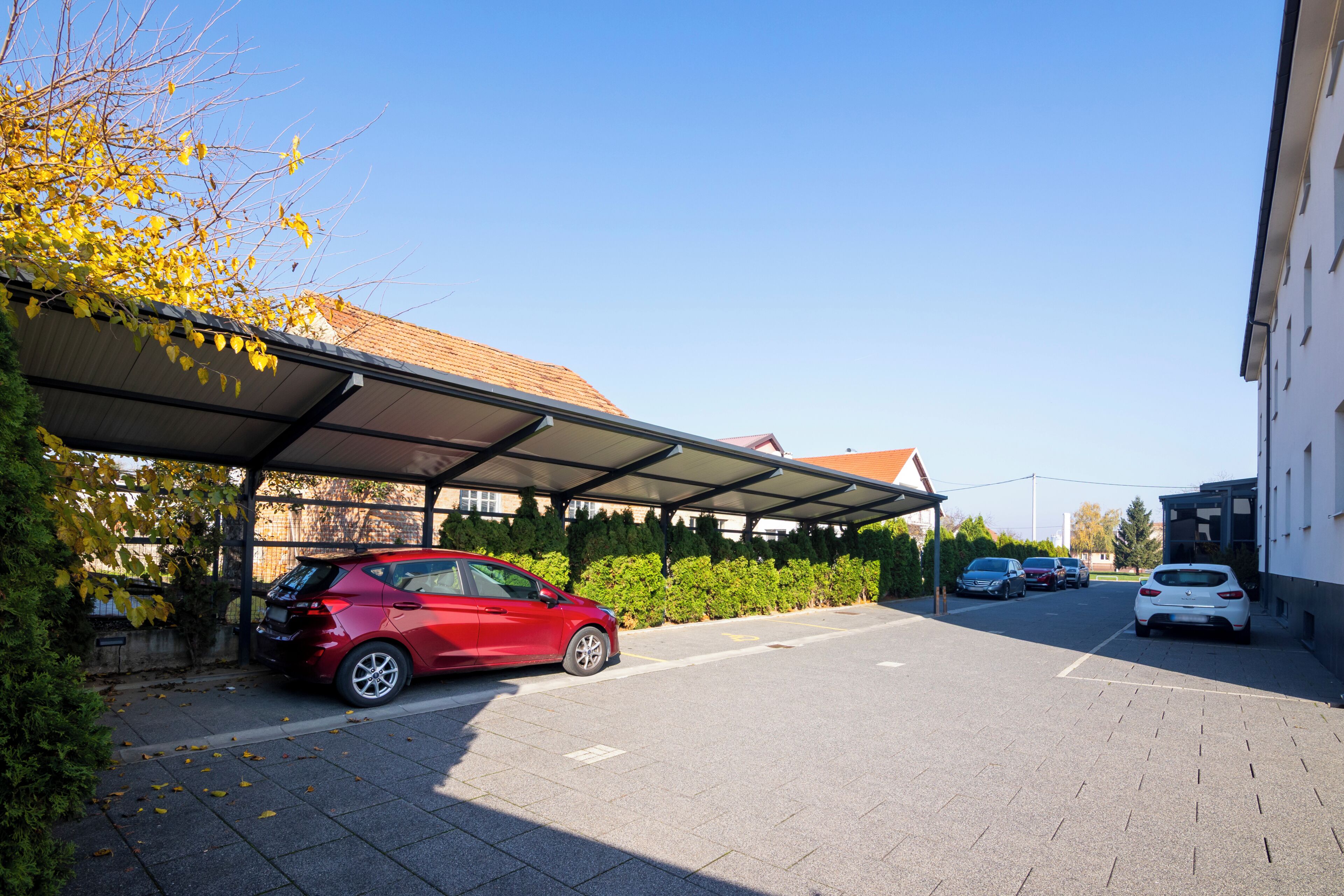 A parking area with a covered carport, a red car, and a white car parked in front of a building with a tiled roof.