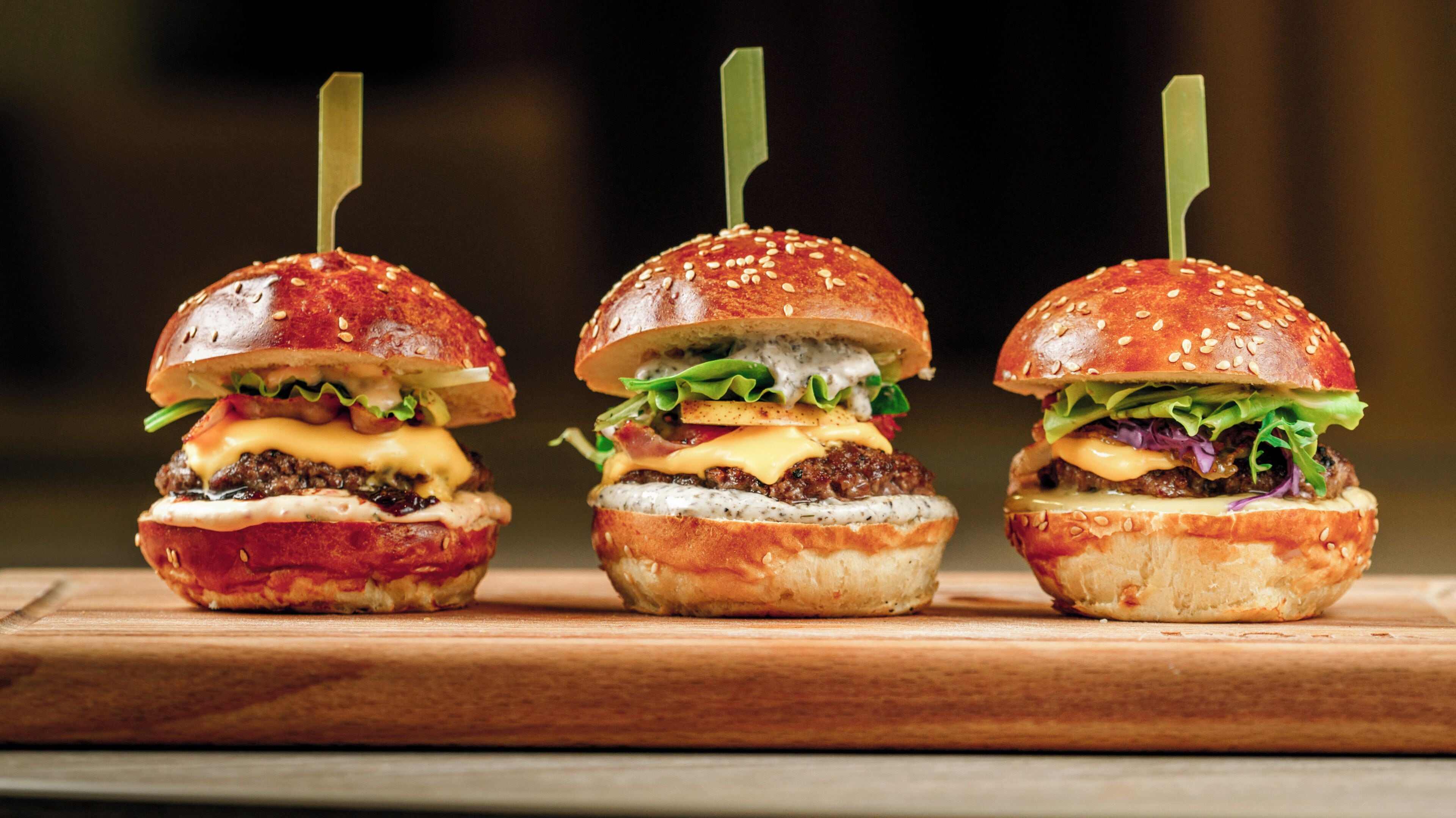 Three burgers with sesame seed buns and toothpicks, placed on a wooden surface.